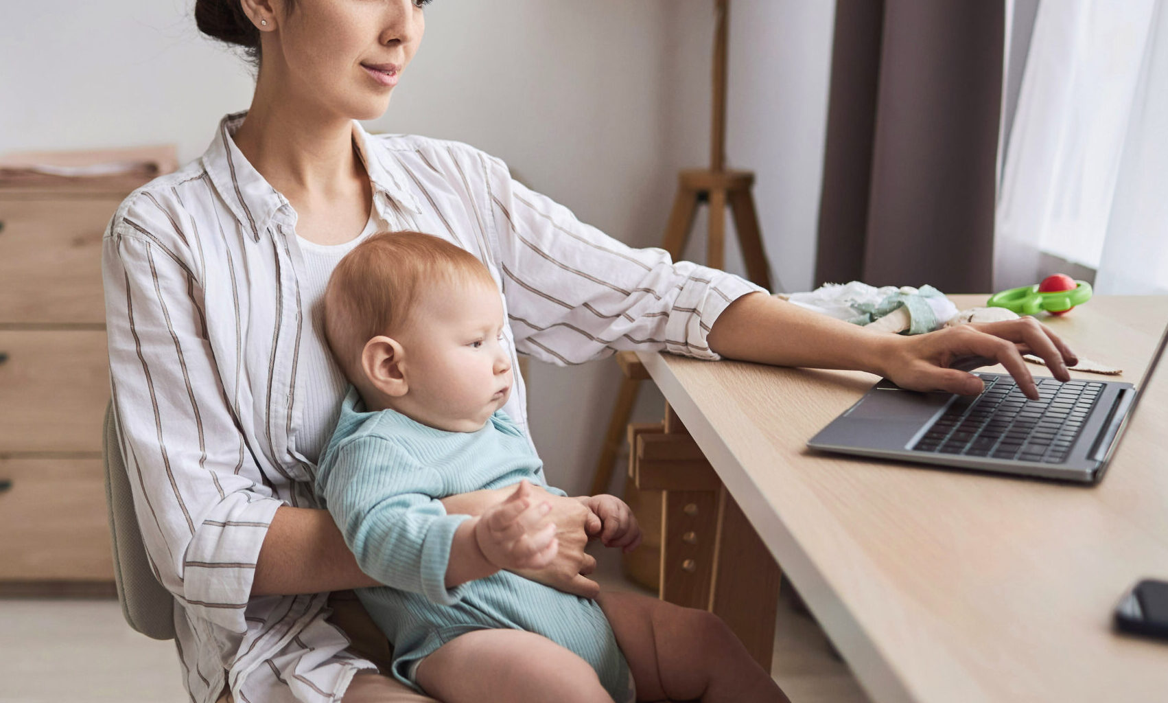 Side view portrait of young mother working from home and holding baby in lap at workplace, copy space